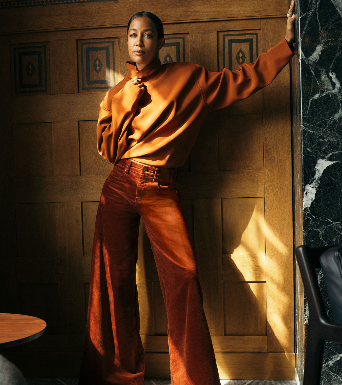 Woman wearing rust-orange wide-leg pants with a matching oversized sweater, standing in a warm, elegant hotel interior with wooden paneling.