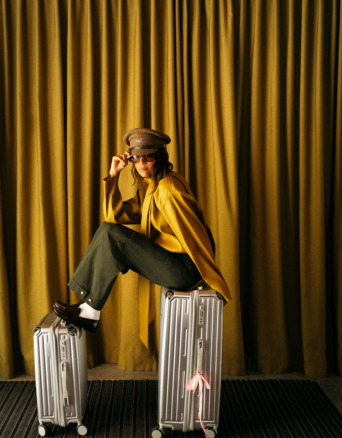 Woman wearing dark wide-leg pants and a mustard yellow jacket, sitting on silver suitcases in front of golden curtains in a stylish hotel interior.
