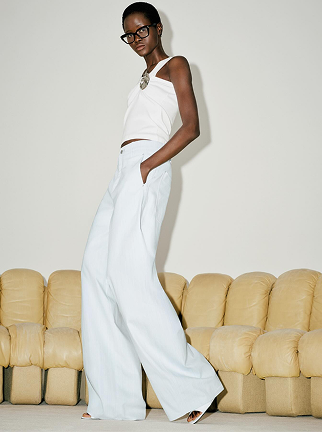 Model wearing white wide-leg pants with a matching asymmetrical sleeveless top, styled with black glasses and statement jewelry. The monochrome look is photographed in front of a cream-colored sofa, highlighting the flowing silhouette and elegant fit.