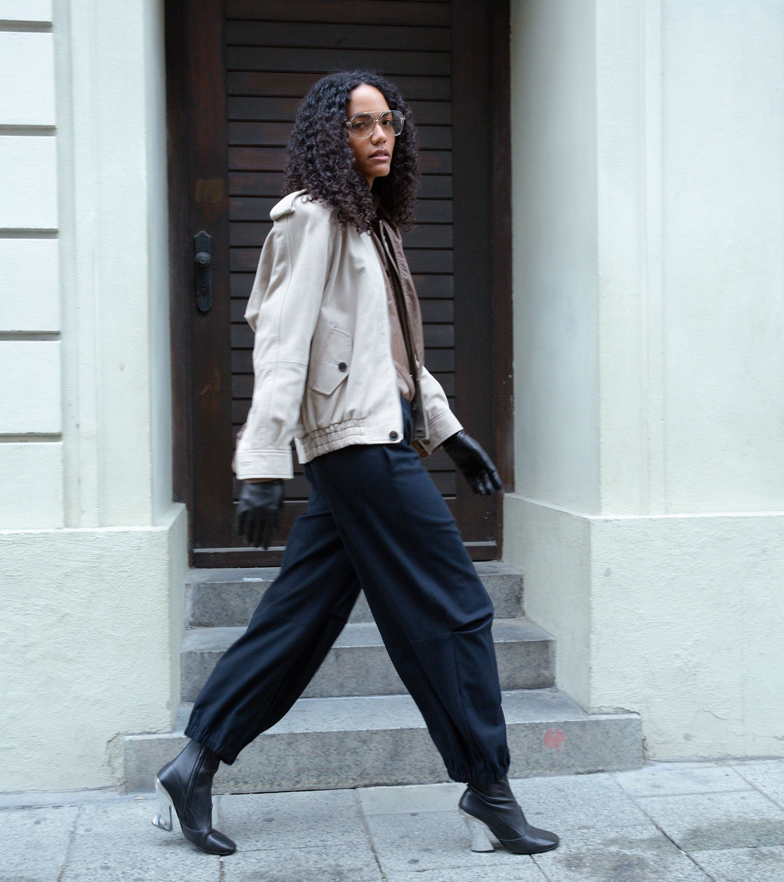 Person with curly hair and glasses walks past steps, wearing a beige jacket, black gloves, wide dark trousers, and heeled boots.