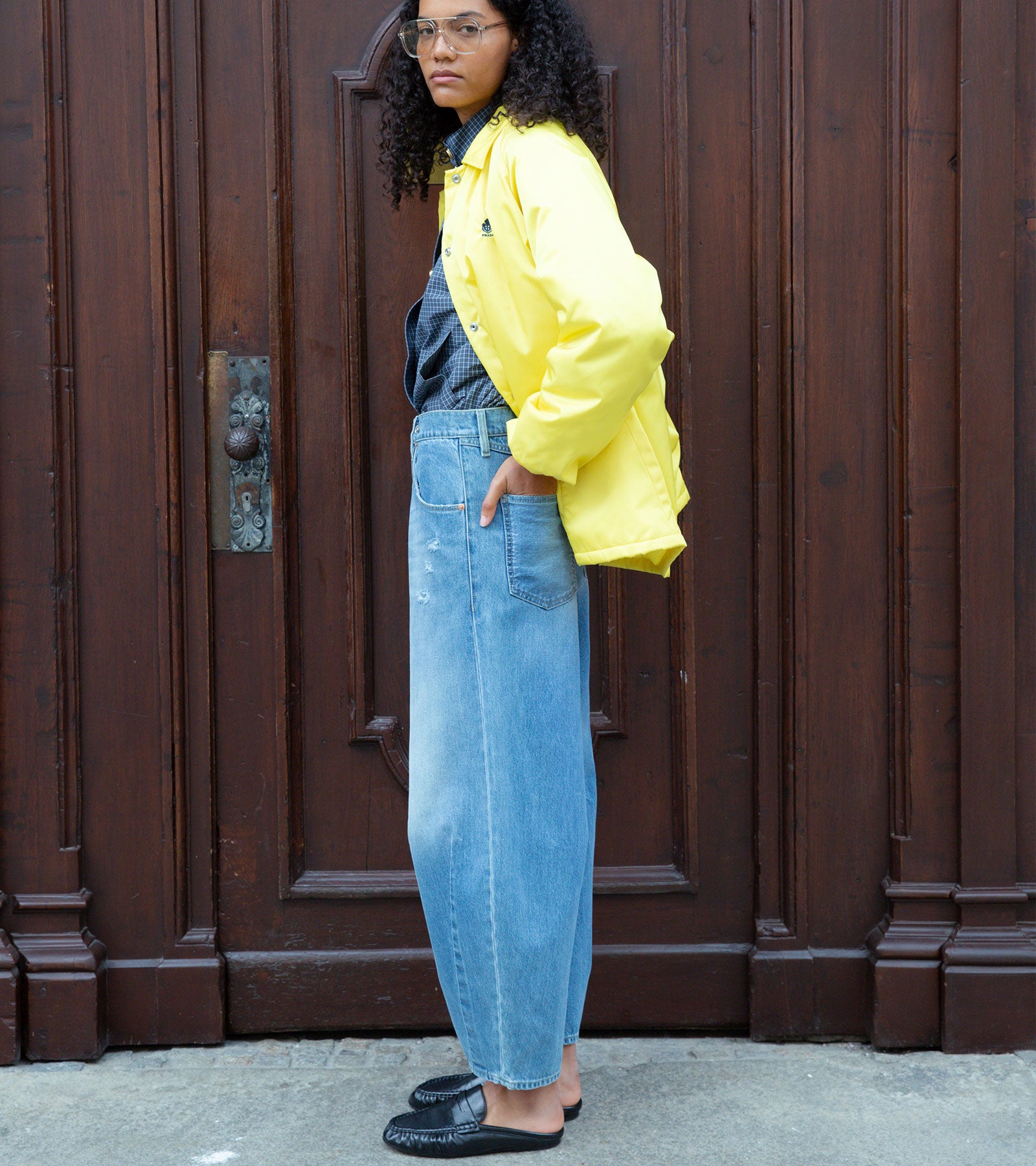 Person with curly hair and glasses poses sideways in a yellow jacket, blue jeans, and black loafers before a dark wooden door.