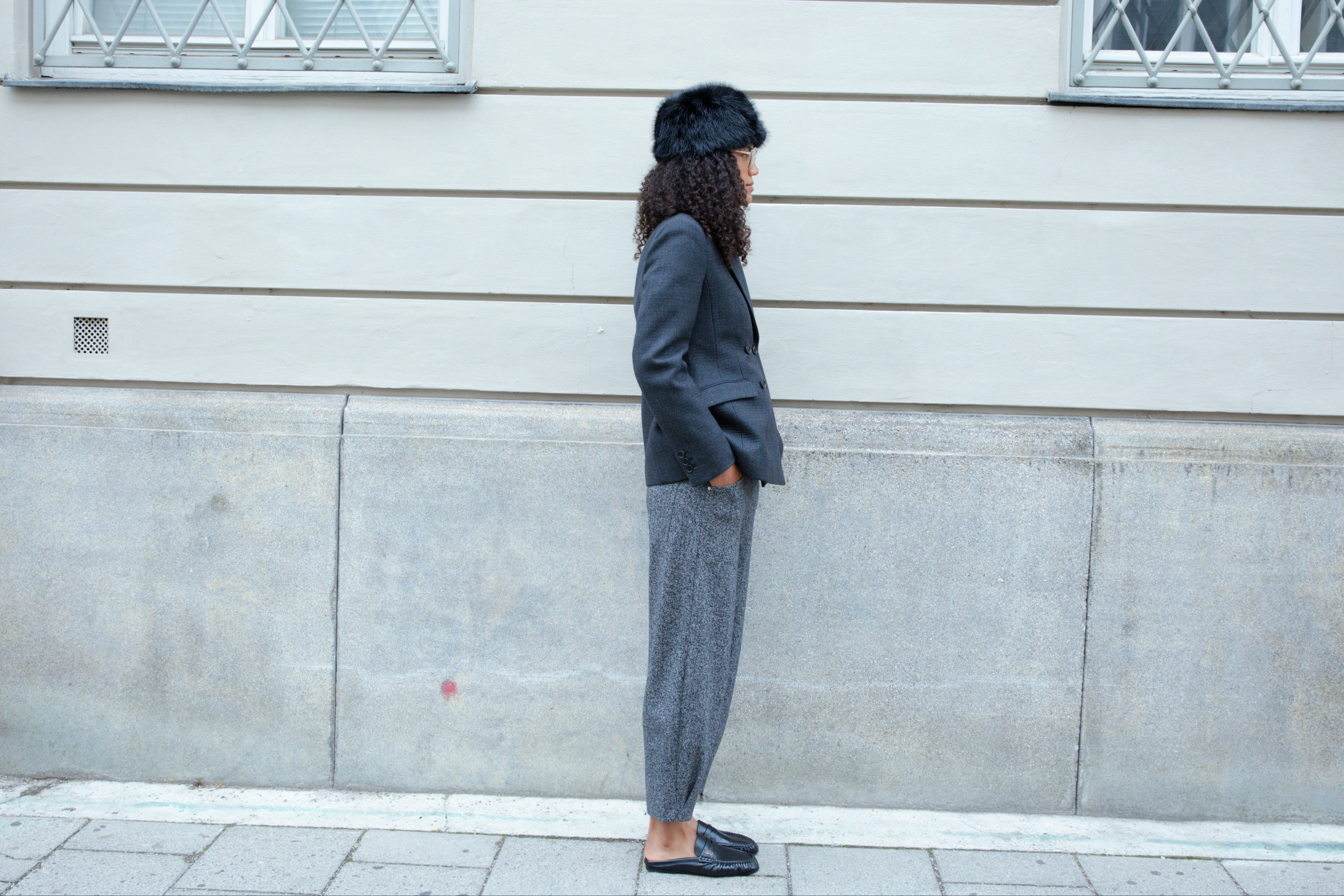 A woman in profile wearing a dark blazer, grey trousers, black flats, and a black fur hat stands against a pale stone building.