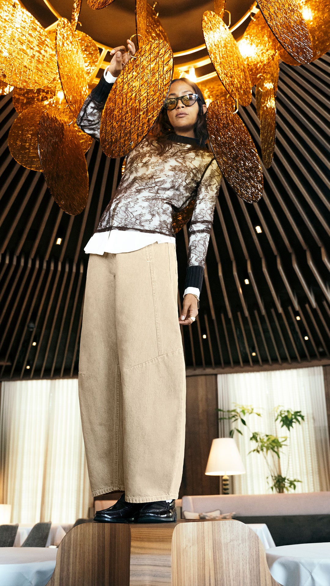 Woman wearing beige wide-leg barrel jeans and a printed top, standing beneath statement golden lights in a modern hotel interior.