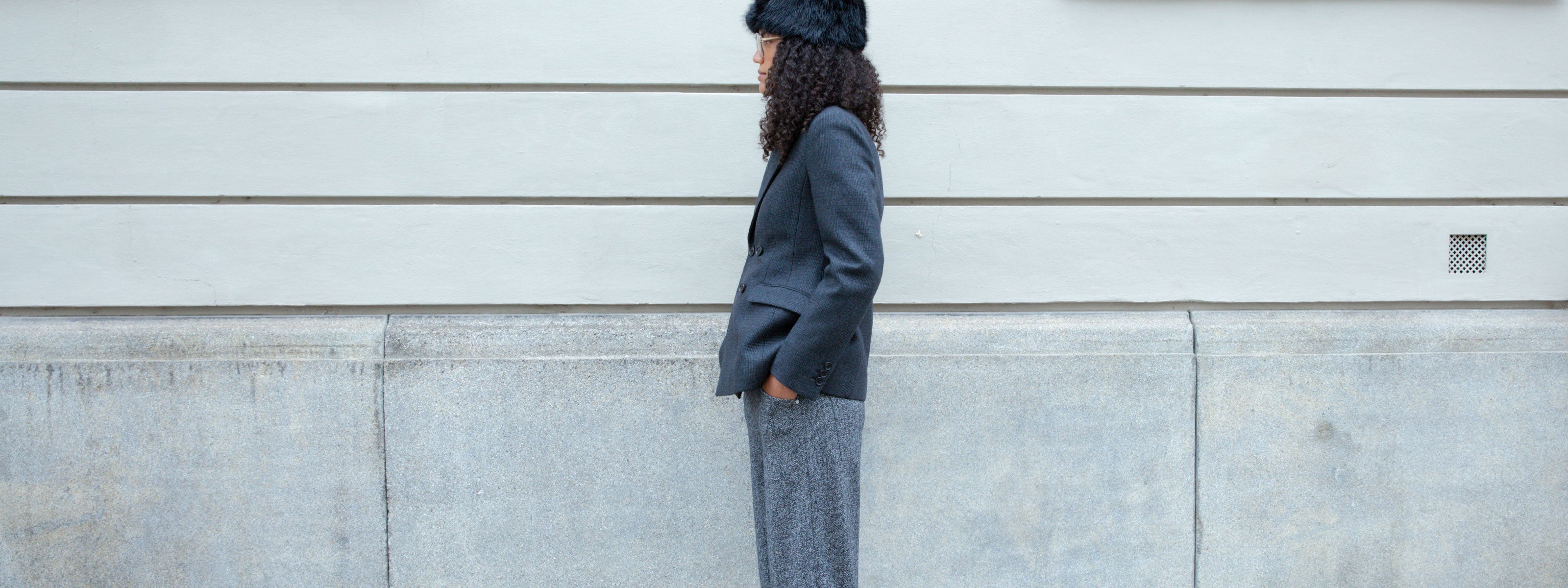 Woman wearing a charcoal blazer and wide-leg trousers standing beside a minimalist stone wall.