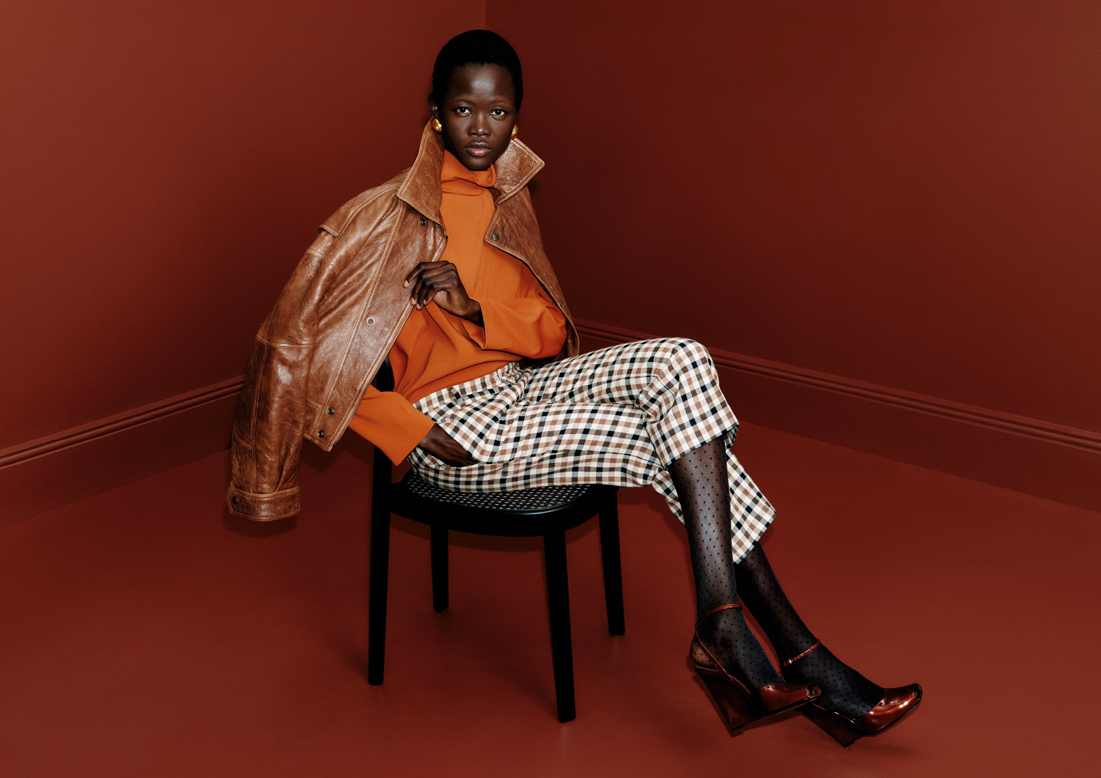 Black woman seated on a chair in an orange blouse, plaid trousers, sheer dotted tights, brown heels, and a leather jacket draped over her shoulders.