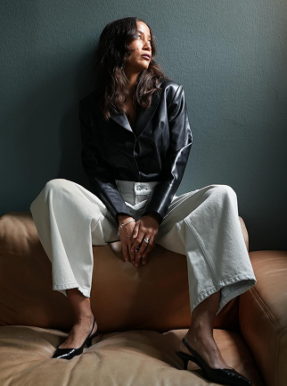 Model wearing a black leather jacket with white barrel-leg jeans and black pointed heels. Seated on a tan leather sofa against a dark wall, the look highlights the relaxed silhouette and modern contrast of monochrome styling.