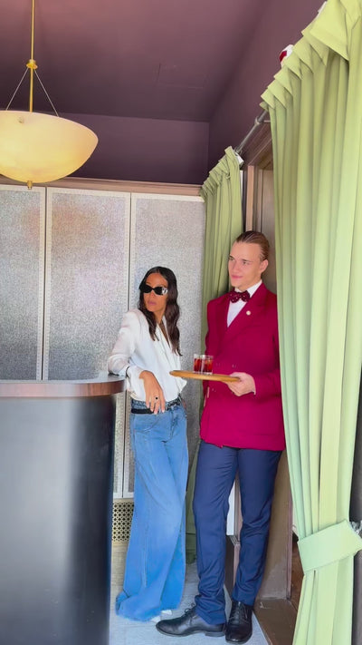 Woman wearing a blue wide-leg jeans and a white blouse standing next to a hotel employee in a red jacket and dark trousers, holding a tray with a drink.